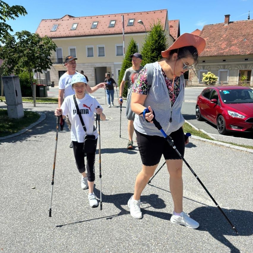 A family is hiking on a road with walking sticks. A red car is parked on the right side of the road.
