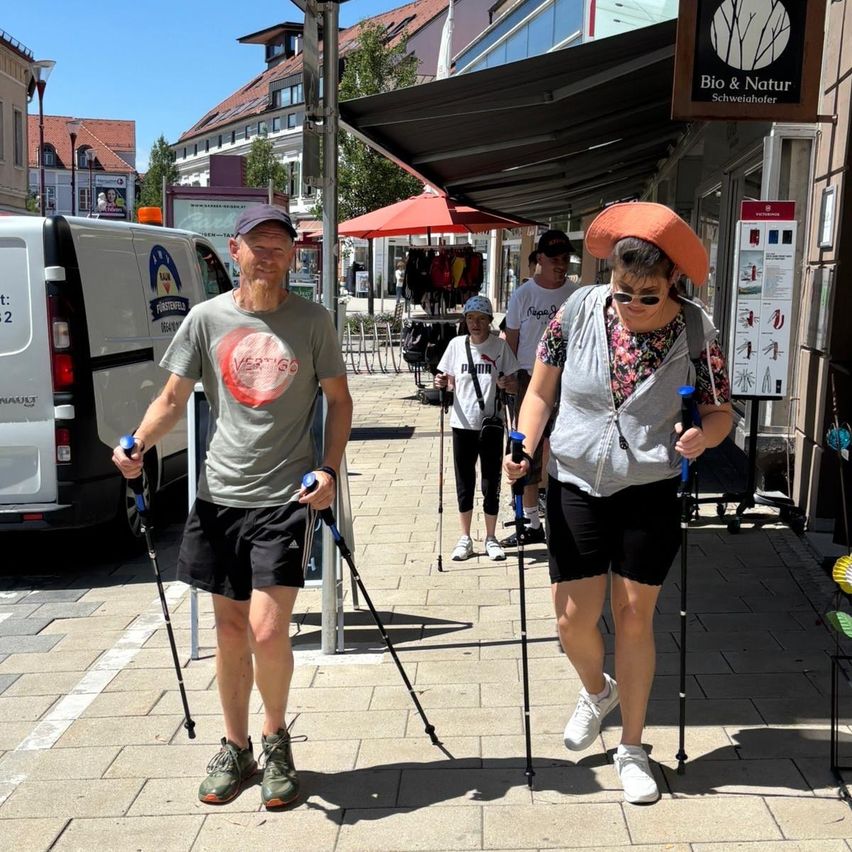 A man and woman, wearing hats and holding trekking poles, are walking on a brick sidewalk in front of a store.