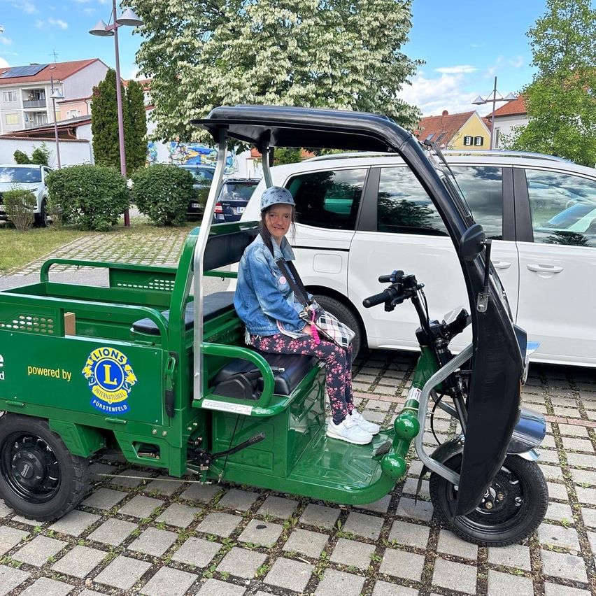 Eine Frau sitzt lächelnd auf einem grünen Elektro-Tuk-Tuk. Es steht auf einer gepflasterten Straße. Der Tuk-Tuk wird von Lions International angetrieben.