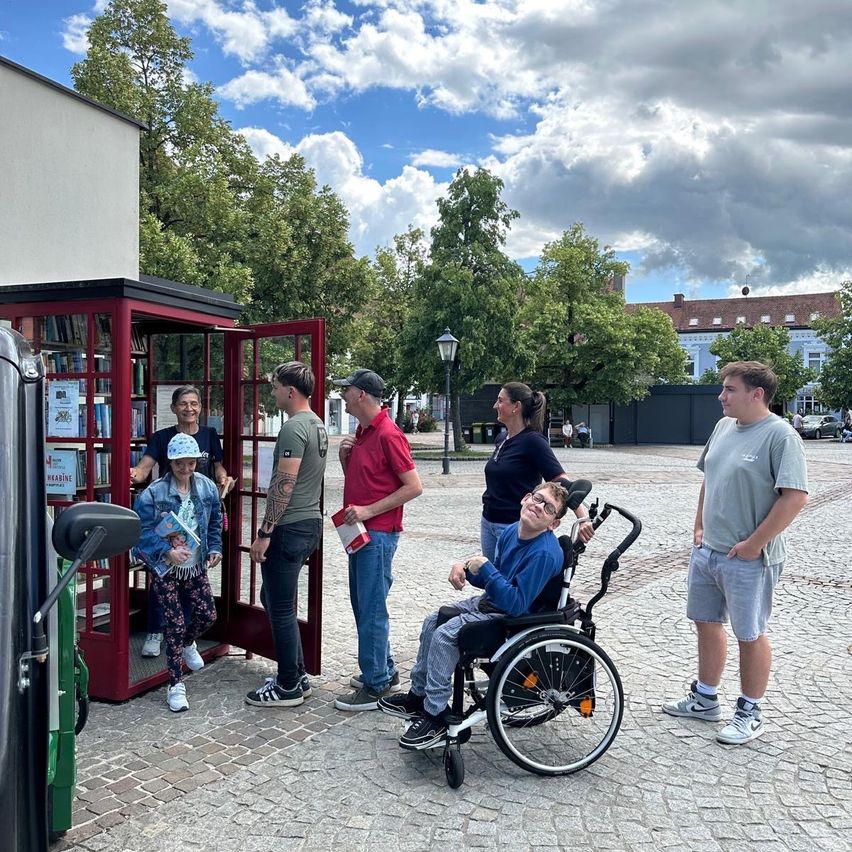 Eine Gruppe von Menschen versammelt sich um eine rote Telefonzelle, in der ein Junge im Rollstuhl sitzt. Eine Frau steht bei der Telefonzelle und hält ein Buch. Sie befinden sich auf einer Kopfsteinpflasterstraße mit Bäumen und Gebäuden im Hintergrund.