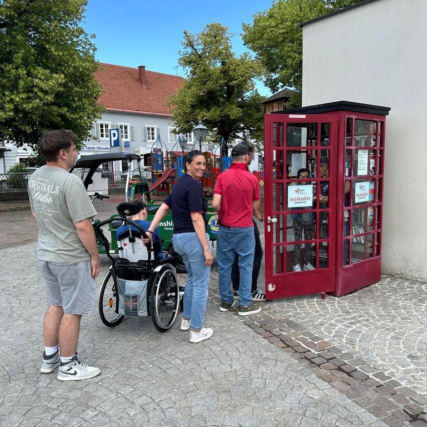 Mehrere Personen stehen um eine rote Telefonzelle auf einem Kopfsteinpflaster. Eine Person im Rollstuhl und eine Frau sind zu sehen, wie sie mit der Telefonzelle interagieren.