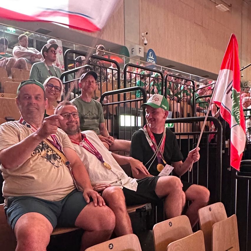 Vier Männer sitzen in einer Arena, lächeln und tragen Medaillen. Sie halten eine Fahne und tragen Hüte. Eine Menschenmenge sitzt im Hintergrund.