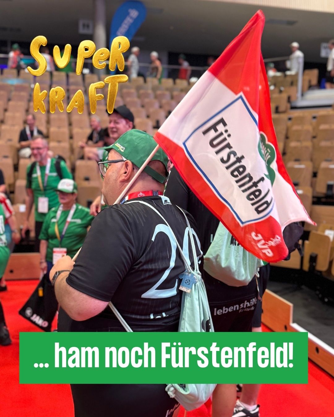 Ein Mann hält eine rot-weiße Flagge mit den Worten 'Fürstenfeld' in einem Stadion. Der Mann trägt einen grünen Hut und eine Brille. Er steht zwischen anderen Leuten. Das Stadion hat leere Sitze.