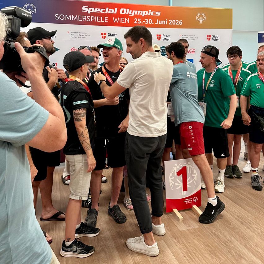 Eine Gruppe von Menschen, darunter auch Athleten, versammelt sich um einen Mann in einem weißen T-Shirt und schwarzen Hosen, möglicherweise bei einer Special Olympics Veranstaltung.