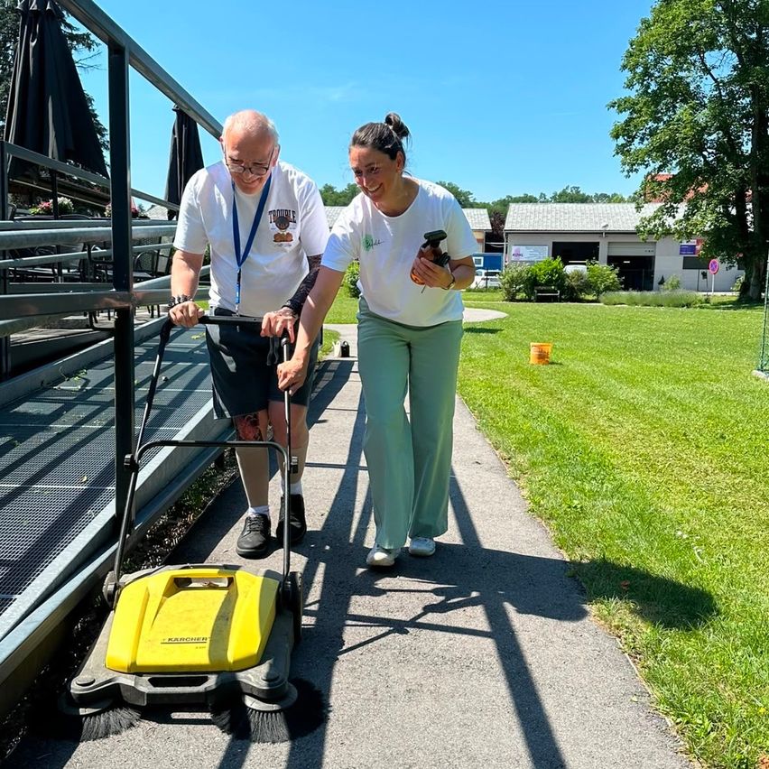 An elderly man and a woman are walking on a path with a Karcher sweeper, smiling in a sunny outdoor setting.