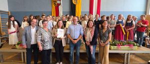 Eine Gruppe von Erwachsenen und Kindern posiert für ein Foto in einem Schulauditorium. Ein Mann in einem blauen Hemd hält ein Zertifikat und eine Frau lächelt. Hinter ihnen hängen drei Flaggen an der Wand.