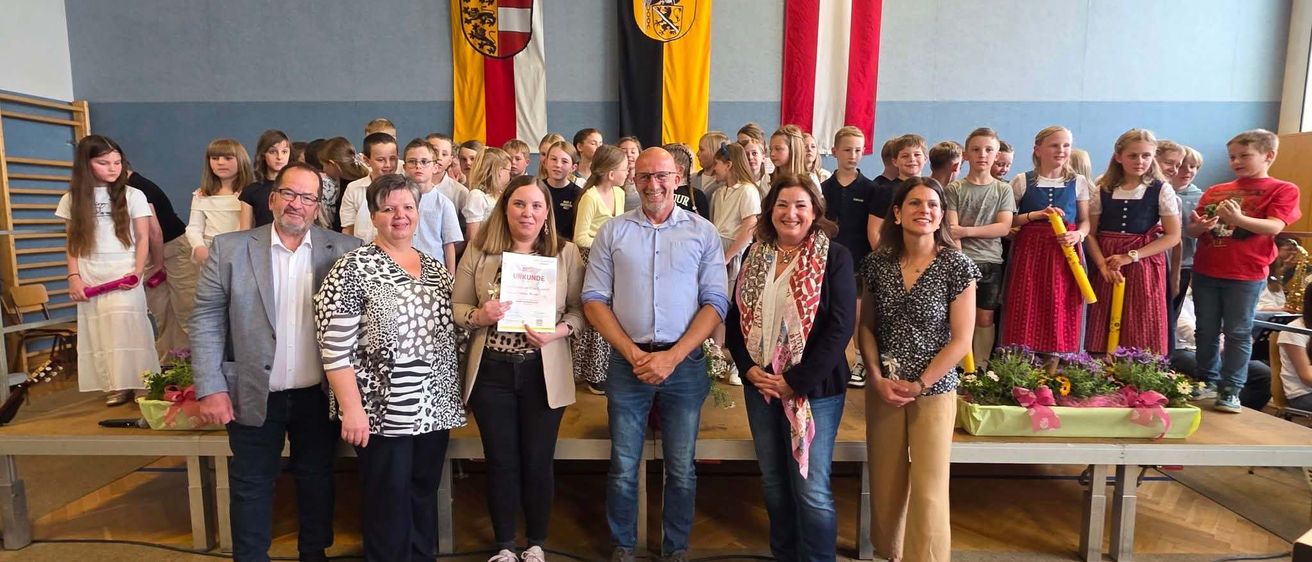 Eine Gruppe von Erwachsenen und Kindern posiert für ein Foto in einem Schulauditorium. Ein Mann in einem blauen Hemd hält ein Zertifikat und eine Frau lächelt. Hinter ihnen hängen drei Flaggen an der Wand.