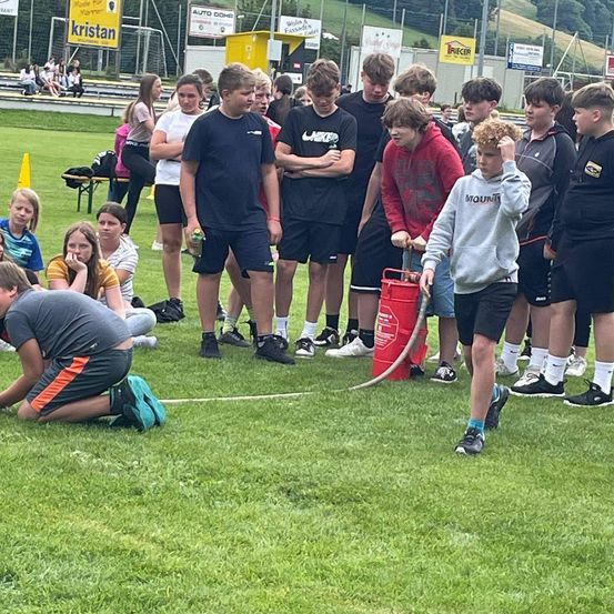 Eine Gruppe von Jungen und Mädchen auf einem Rasenfeld, wahrscheinlich für eine Sportveranstaltung. Ein Junge in einer grauen Kapuzenjacke hält ein rotes Objekt und scheint mit dem Jungen vorne zu sprechen.