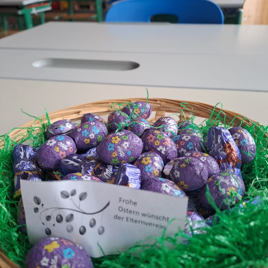 Ein Korb mit lila Schokoladeneiern mit Blumen steht auf einem Tisch mit einem Zettel, der Frohe Ostern wünscht der Elternverein.
