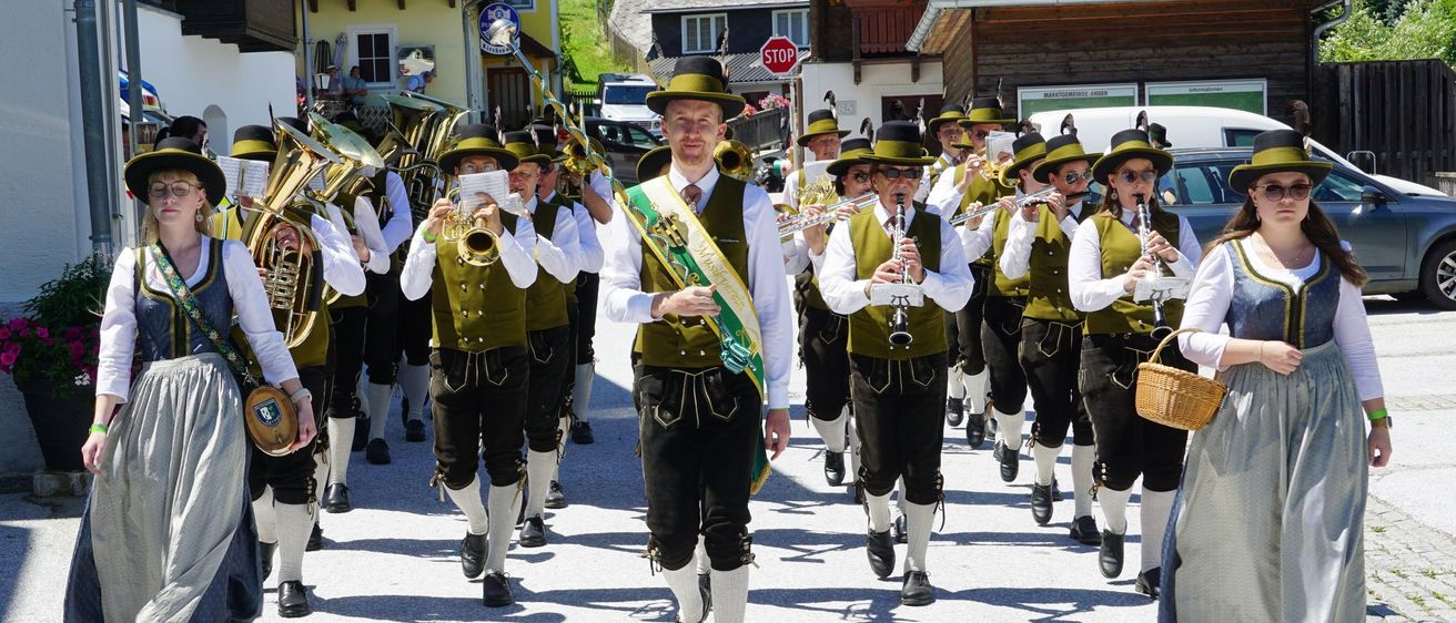 Eine Blaskapelle in traditioneller Kleidung tritt bei einem Umzug auf. Sie spielen Blech- und Holzblasinstrumente, einige Mitglieder tragen Tuba. Ein Mann führt die Kapelle mit einer Schärpe.