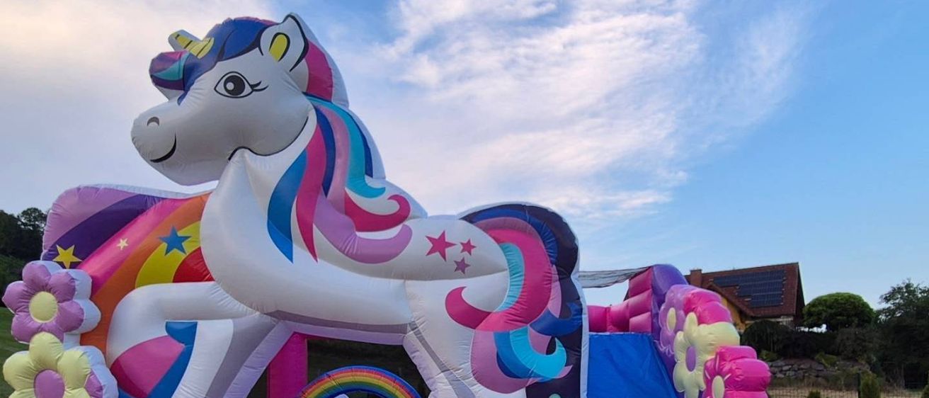Ein buntes aufblasbares Einhorn-Sprunghaus mit Rutsche und Blumen, auf einer Rasenfläche unter einem teilweise bewölkten Himmel aufgestellt.