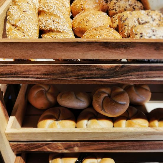 Ein Holztablett hält eine Vielzahl von Brötchen und Buns, darunter sesambestreute und brezelförmige. Sie sind ordentlich gereiht.