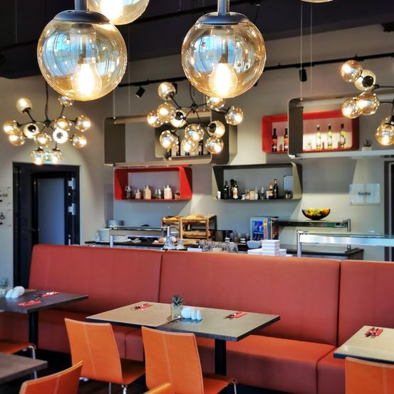 Ein modernes Restaurant mit roten Sofas und orangefarbenen Stühlen, Pendelleuchten und Regalen mit Flaschen. Der Küchenbereich hat eine Glastheke, eine Obstschale und verschiedene Küchenutensilien.