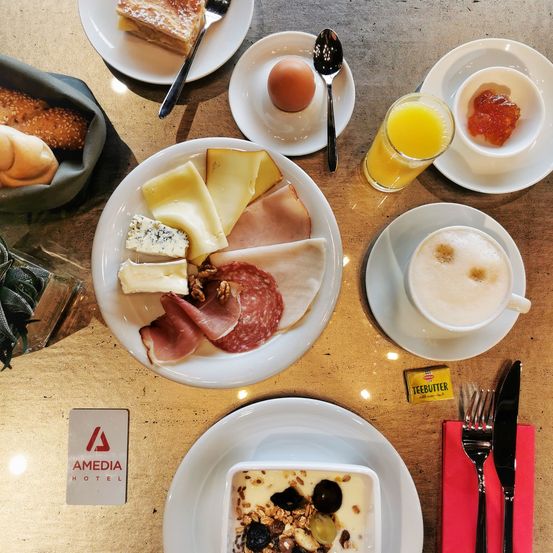 Ein Frühstücksbuffet mit verschiedenen Käsesorten, Fleisch und einer Tasse Kaffee auf einem Tisch. Andere Gegenstände sind Eier, Brot und ein Glas Orangensaft.