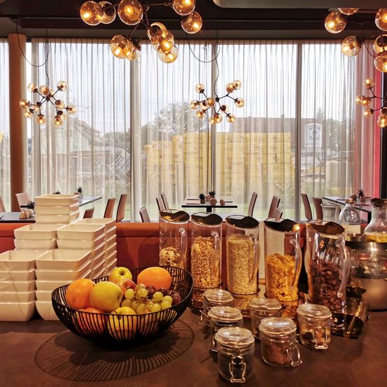 Ein Frühstücksbereich mit einem Tisch, auf dem Obst, Müsli und Getränke stehen. Dahinter bietet ein großes Fenster mit Vorhängen einen Blick auf einen Außenbereich.