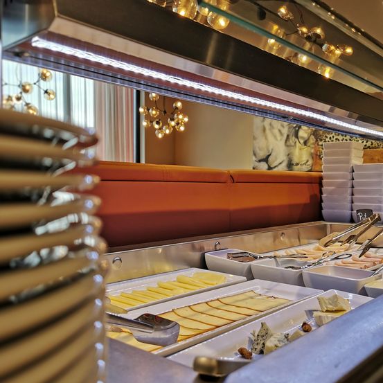 Ein Buffettisch mit verschiedenen Käsesorten, Fleisch und Nüssen, beleuchtet von hellen Lichtern. Der Tisch ist mit weißen Tellern bedeckt, und eine Kronleuchter hängt darüber.