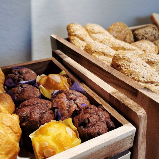 Zwei Holzkisten enthalten eine Auswahl an Gebäck, darunter Muffins, Croissants und Brot mit Sesamkörnern. Die Muffins sind dunkel und scheinen schokoladig zu sein.