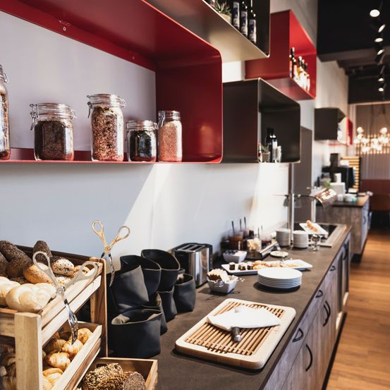 Ein Buffet-Tresen mit Brot und Gebäck auf einem Holzregal. Verschiedene Gegenstände und Gläser befinden sich auf den roten Regalen. Teller, Schüsseln und Tassen stehen auf dem Tresen.
