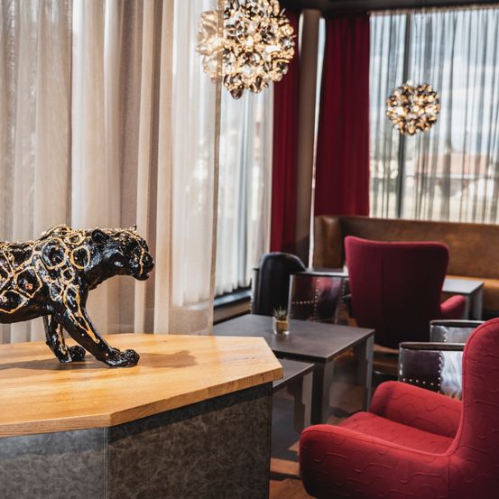 Ein Raum mit einem hölzernen Tisch, auf dem eine dekorative schwarze Pantherstatue steht. Es gibt rote Stühle, eine Couch und ein Fenster mit Vorhängen. Zwei Kronleuchter hängen von der Decke.