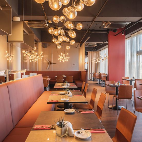 Ein Innenraumblick eines modernen Restaurants mit großen hängenden Lichtkörpern, hölzernen Sitzbänken und Tischen. Der Raum ist gut beleuchtet, mit roten Akzenten und weißen Vorhängen.