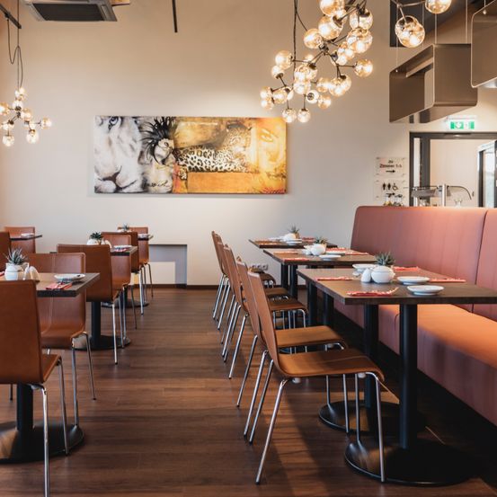 Ein Restaurant mit braunen Lederstühlen um Tische, einem großen Löwenbild an der Wand und Pendelleuchten an der Decke.