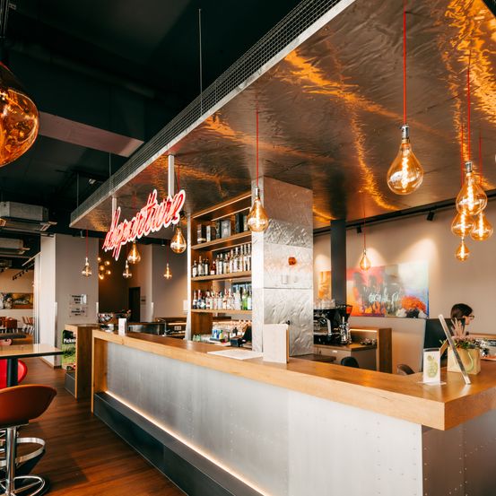 Ein Innenansicht eines Cafés mit einer Holztheke, Regalen mit Schnapsflaschen, einem roten Neonschild mit der Aufschrift 'Adventure' und mehreren hängenden Glühbirnen. Eine Person sitzt an einem Tisch, und verschiedene Möbelstücke sind zu sehen.