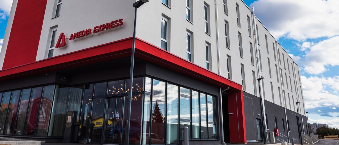 Das moderne Hotelgebäude, gekennzeichnet als Amedia Express, verfügt über eine weiße Fassade mit roten Akzenten, große Glasfenster und einen gläsernen Eingang. Der Himmel ist blau mit Wolken.