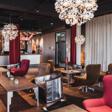 Ein elegantes Wohnzimmer mit verschiedenen Stühlen, Tischen und Kronleuchtern. Ein Feuerlöscher ist an der Wand montiert, und eine Theke mit einem Neonschild ist in der Ferne.