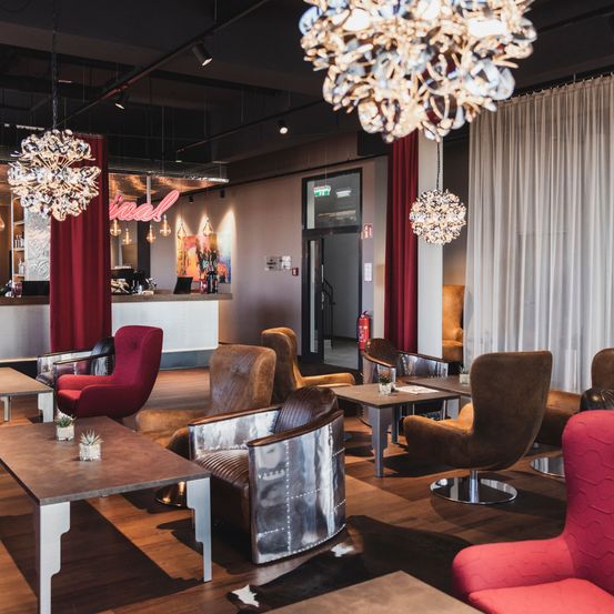 Ein elegantes Wohnzimmer mit verschiedenen Stühlen, Tischen und Kronleuchtern. Ein Feuerlöscher ist an der Wand montiert, und eine Theke mit einem Neonschild ist in der Ferne.