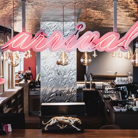 Ein Restaurant-Interieur zeigt ein rosa Neonschild mit der Aufschrift 'animal'. Eine goldene Jaguar-Statue steht auf einem Holztisch. Dahinter befindet sich eine Theke mit einer Kaffeemaschine und anderen Geräten. Regale, ein Schrank und eine Glaswand sind sichtbar.