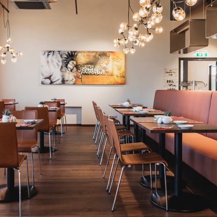 Ein leeres Restaurant mit Lederstühlen und runden Tischen, einem großen Leopardenbild an der Wand und von der Decke hängenden Kronleuchtern.