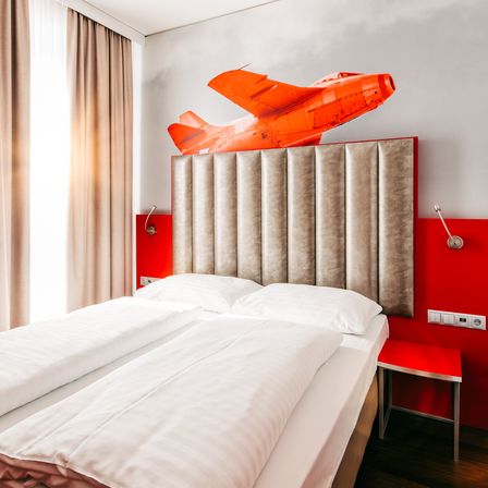 Ein Schlafzimmer mit einem Doppelbett, weißen Laken und Kissen. Die Wand ist weiß mit einem orangefarbenen Flugzeugdesign. Ein roter Nachttisch und eine Lampe befinden sich auf der rechten Seite.