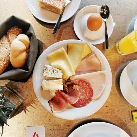 Ein Frühstückstisch zeigt einen großen Teller mit verschiedenen Käsesorten, Fleisch und Brot. Daneben steht ein Teller mit einem Ei und einem Löffel. Ein Korb mit Brot und ein Glas Orangensaft sind ebenfalls vorhanden.