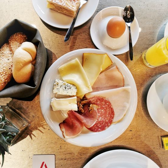 Ein Frühstückstisch zeigt einen großen Teller mit verschiedenen Käsesorten, Fleisch und Brot. Daneben steht ein Teller mit einem Ei und einem Löffel. Ein Korb mit Brot und ein Glas Orangensaft sind ebenfalls vorhanden.