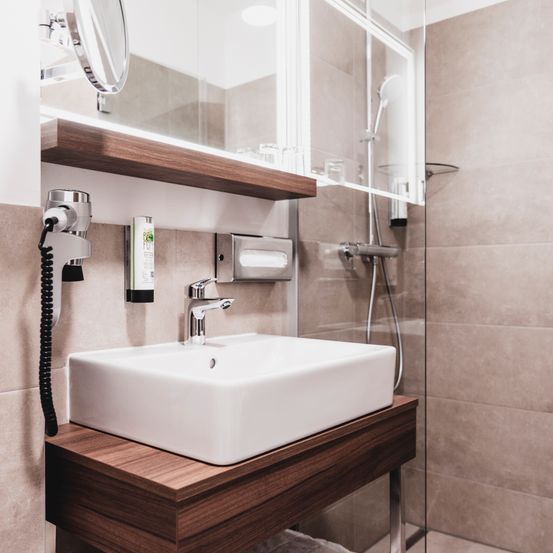 Ein modernes Badezimmer mit einem weißen Waschbecken, einem Holzschrank und einer Glasdusche. Der Schrank hat einen Spiegel, einen Wasserhahn und einen Föhn. Die Dusche hat einen Duschkopf und einen Handtuchhalter.