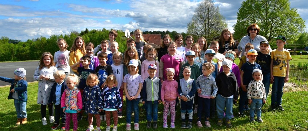 Eine große Gruppe von Kindern und Erwachsenen, die auf einer Grasfläche zusammenstehen, möglicherweise für ein Gruppenfoto, mit bewölktem Himmel und Bäumen im Hintergrund.