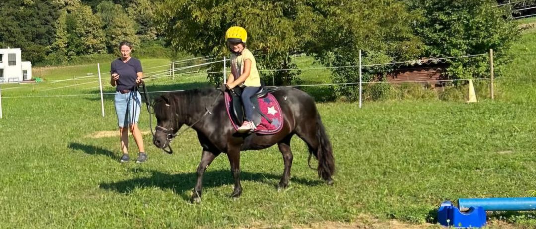 Ein junges Kind reitet auf einem kleinen schwarzen Pferd auf einer Wiese mit einem Mann, der am Halfter zieht. Im Hintergrund steht ein weißes Haus und Bäume.
