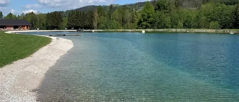 Ein ruhiger See mit klarem Wasser und einem Schotterufer. Grüner Rasen und Bäume umgeben das Gebiet. Ein kleiner Steg ragt in den See hinein.