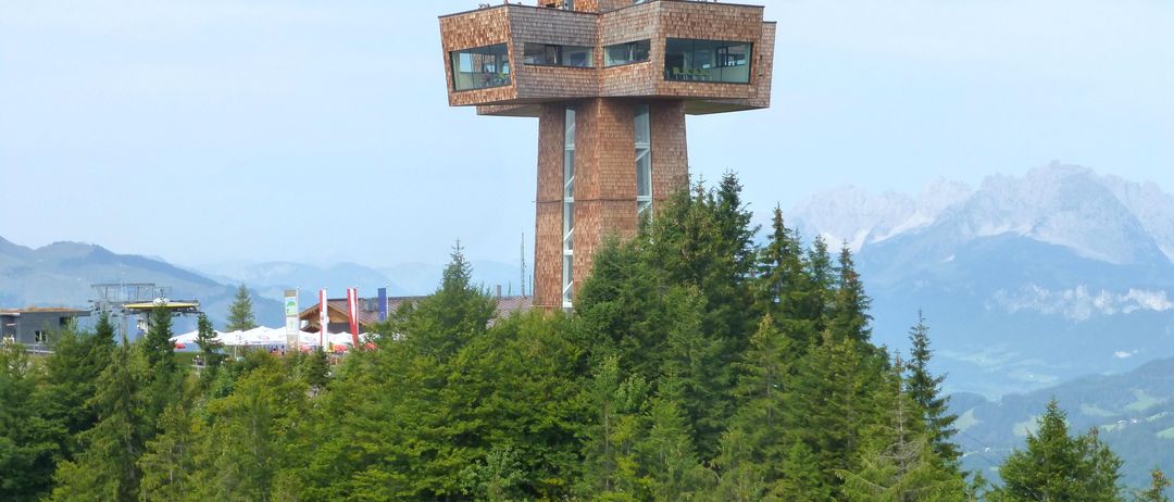 Ein hoher hölzerner Turm in Form eines Kreuzes auf einem Berggipfel, umgeben von Bäumen und Bergen.
