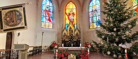 Ein Kirchenaltar, geschmückt mit Blumen und Kerzen. Der Altar hat eine Statue und einen Weihnachtsbaum davor. Die Kirche hat Buntglasfenster.