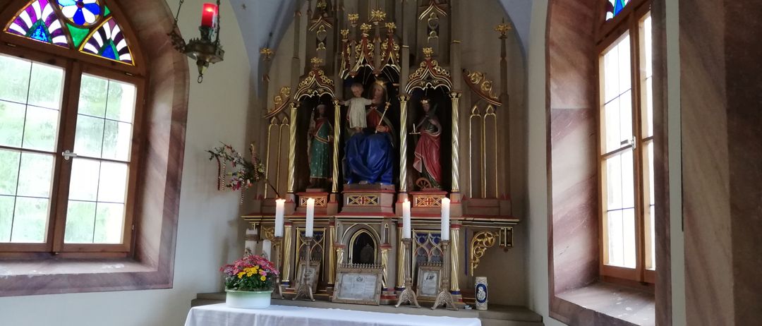 Ein Altar in einer Kirche mit Statuen und Kerzen. Der Altar hat eine weiße Tischdecke. Die Wände sind mit aufwendigen Designs verziert. An den Seiten befinden sich Buntglasfenster.