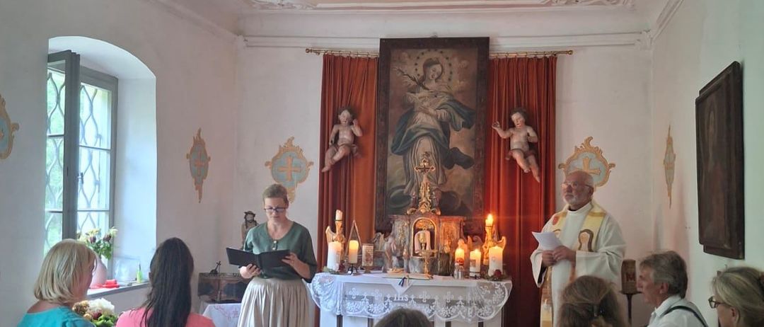 Eine Gruppe von Menschen versammelt sich in einer Kapelle. Eine Frau liest aus einem Buch, während ein Priester aus einem Papier liest. Vor ihnen steht ein Altar mit Kerzen und einem religiösen Gemälde.