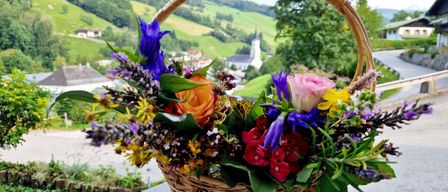 Ein Korb aus Weide mit einem farbenfrohen Blumenstrauß steht auf einer Steinbank mit einer malerischen Kulisse aus Hügeln und einer Dorfkirche im Hintergrund.