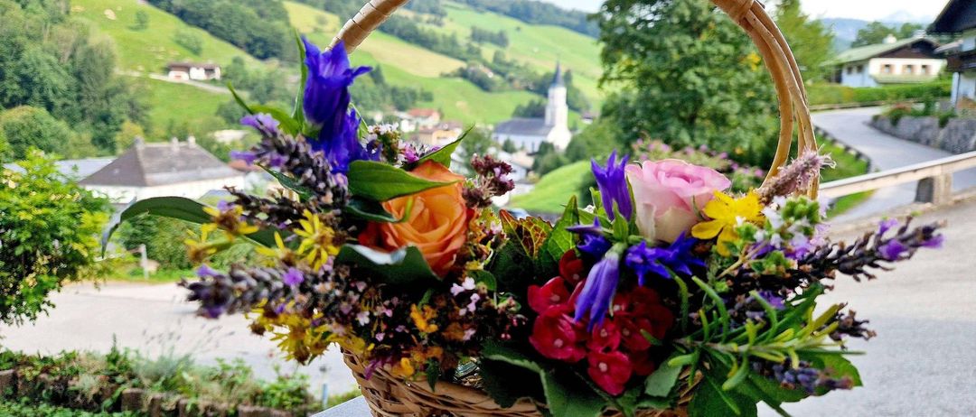 Ein Korb aus Weide mit einem farbenfrohen Blumenstrauß steht auf einer Steinbank mit einer malerischen Kulisse aus Hügeln und einer Dorfkirche im Hintergrund.