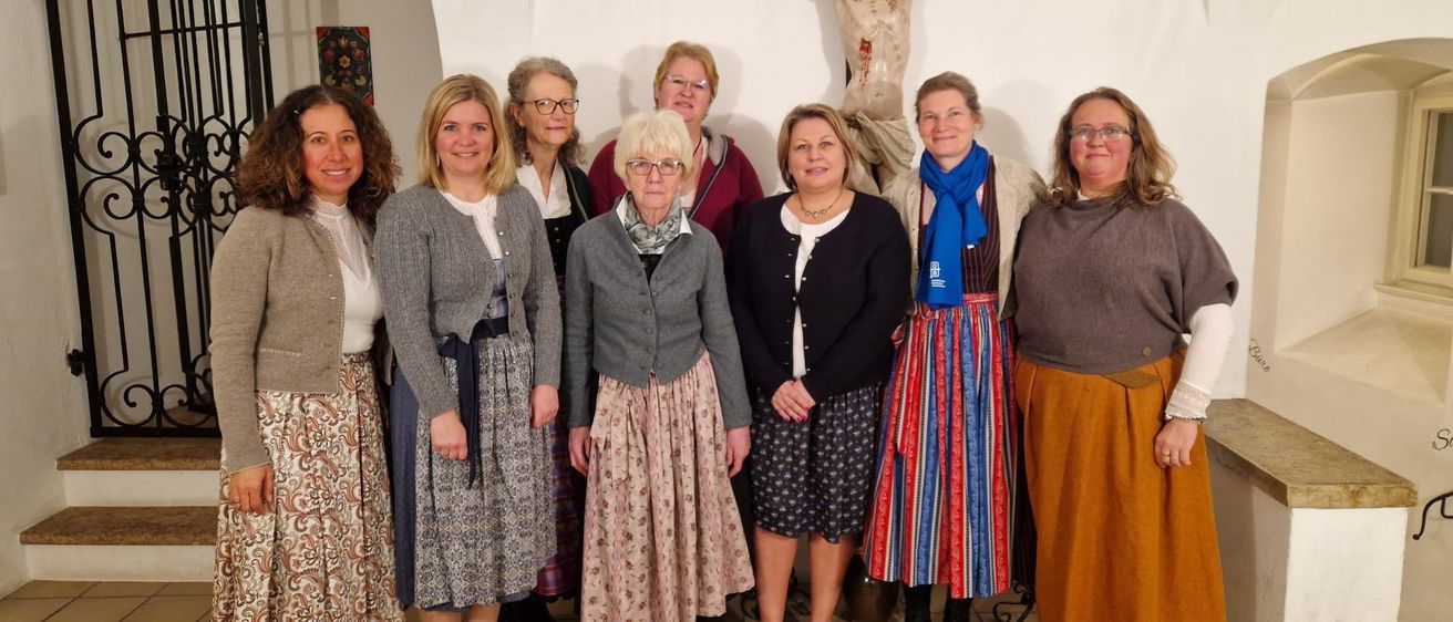 Eine Gruppe von Frauen steht vor einer Kirche, trägt Kleider und lächelt. Sie scheinen für ein Foto zu posieren. Ein Kreuz ist an der Wand hinter ihnen angebracht.