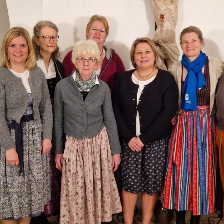 Eine Gruppe von Frauen steht vor einer Kirche, trägt Kleider und lächelt. Sie scheinen für ein Foto zu posieren. Ein Kreuz ist an der Wand hinter ihnen angebracht.