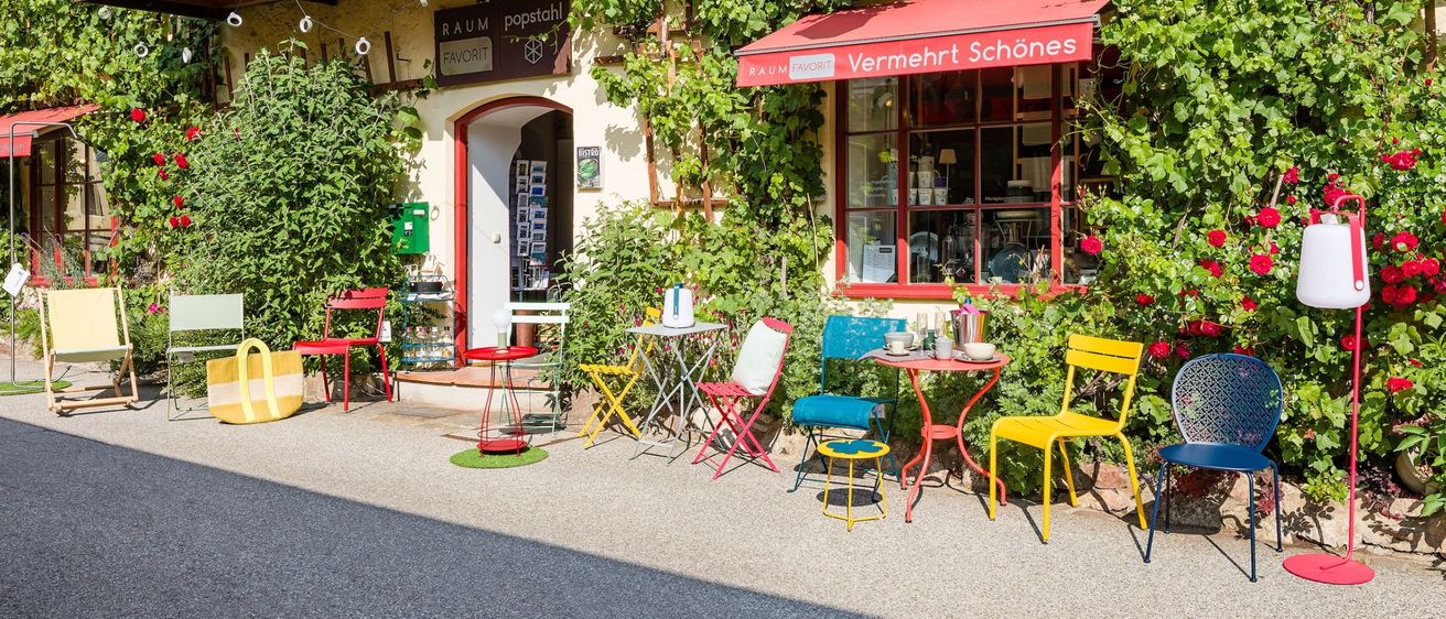Ein entzückendes Geschäft mit einem roten Vordach namens 'Vermehrt Schönes', verfügt über einen Balkon mit Geländern und üppigen grünen Ranken. Draußen sind farbenfrohe Stühle und Tische arrangiert.