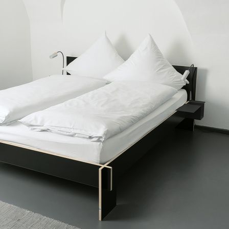 Ein modernes Bett mit schwarzem Rahmen und weißer Bettwäsche steht an einer weißen Wand. Zwei Lampen sind am Kopfteil montiert.