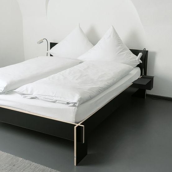 Ein modernes Bett mit schwarzem Rahmen und weißer Bettwäsche steht an einer weißen Wand. Zwei Lampen sind am Kopfteil montiert.