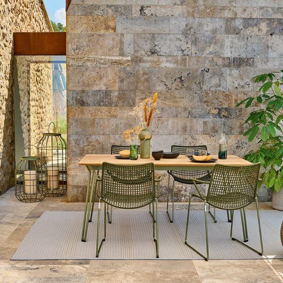 Eine Outdoor-Essplatz-Einrichtung mit einem Holztisch und vier grünen Stühlen auf einem Steinboden. Ein Topf mit Pflanzen und Dekorationsartikeln stehen auf dem Tisch.